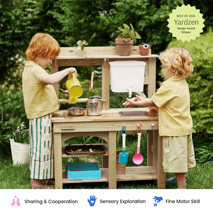 Two children playing with a wooden pretend play kitchen outdoors on a grassy lawn. The wooden play kitchen has a sink, pots, pans, hanging utensils, and shelves.
