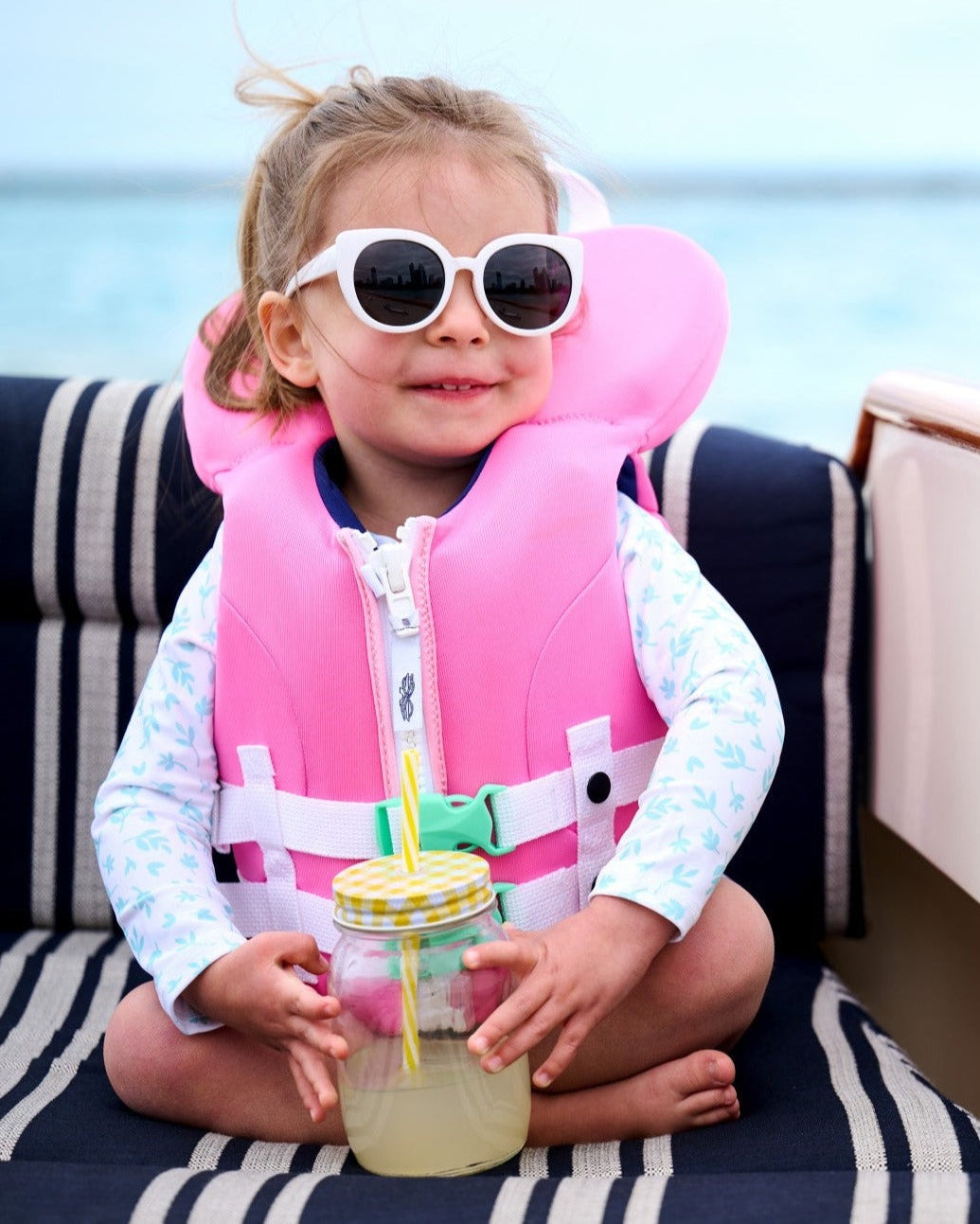 Child wearing a pink life jacket and sunglasses, sitting on a striped surface with a drink.